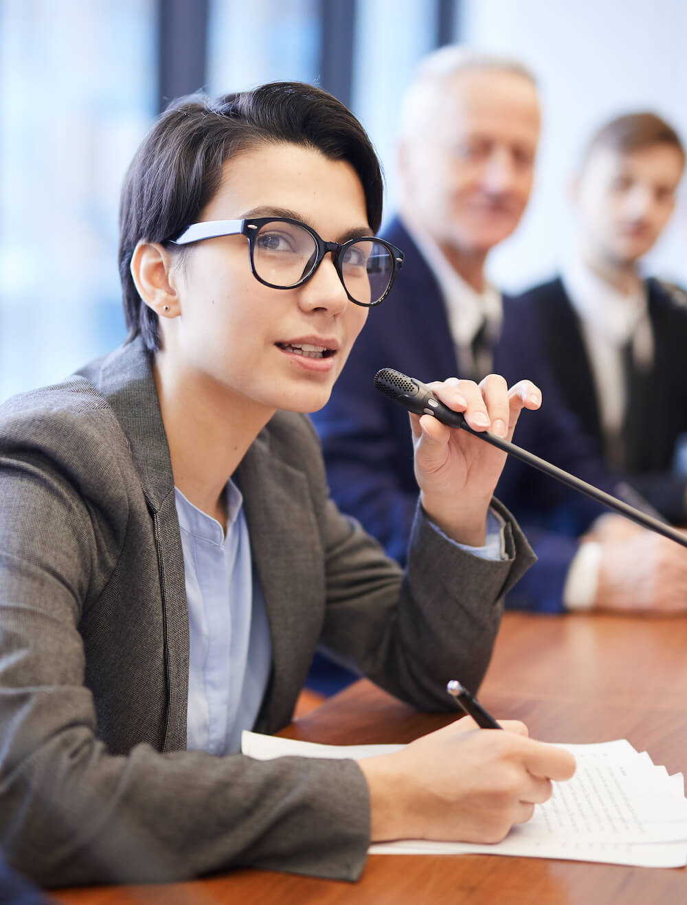 Portrait of young businesswoman speaking to microphone during conference and taking notes while sitting in row with colleagues , copy space